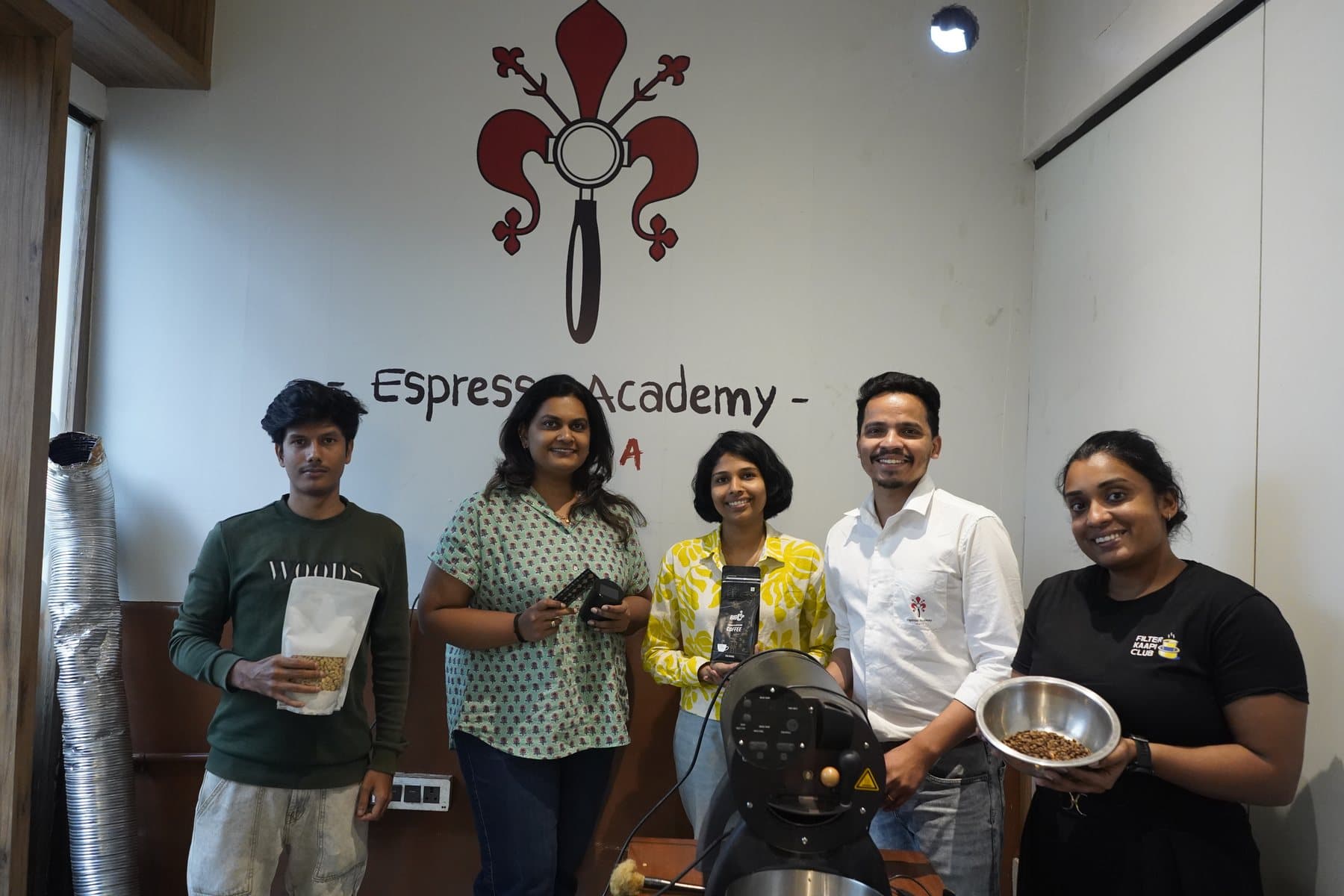 Five people standing under the Espresso Academy fleur-de-lis painted on the campus wall, holding green beans, roasted beans and a bag of coffee, with the drum roaster in front of them.